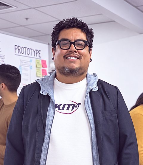 Strike Heredia smiling, wearing glasses, a jacket and a white T-shirt, at an innovation workshop with prototype boards and sticky notes